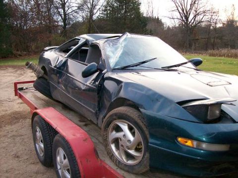 1996 Ford Probe - A heavily damaged dark green coupe sits on a trailer, its roof caved in and windshield shattered.