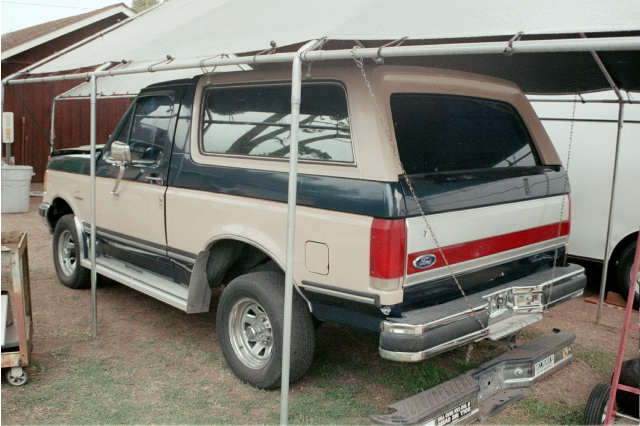 Tan and dark blue Ford Bronco SUV, rear three-quarter view, parked under a carport with parts on the ground