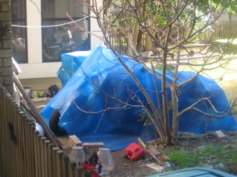 1986 Ford Bronco - Vehicle covered in blue bubble wrap, parked outdoors near a building and fence.
