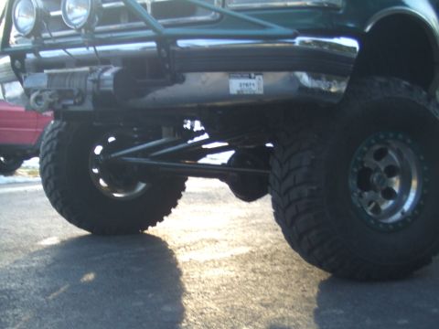 1996 Ford Bronco - Close-up of the front suspension and large tires on a 1996 Ford Bronco, with another vehicle in the background.