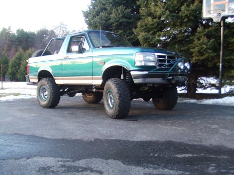 1996 Ford Bronco - Three-quarter front view of a lifted teal green and tan two-tone Ford Bronco parked on a driveway with the hardtop removed and roll bar vis…