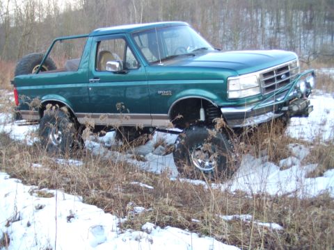 1996 Ford Bronco - Three-quarter front view of a lifted teal green and tan two-tone Ford Bronco parked in snowy brush with the hardtop removed, roll bar in pl…