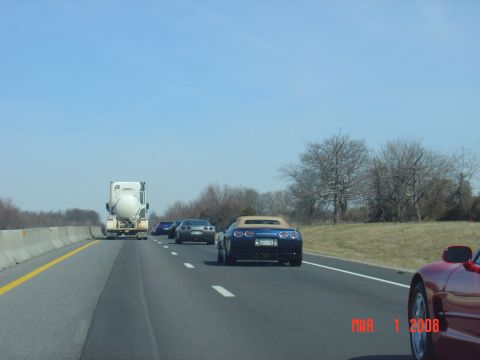 1986 Mazda RX-7 - A blue convertible sports car is seen from behind on a highway, with other vehicles and a tanker truck in the distance.