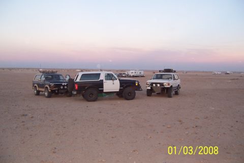 1990 Ford Bronco - Three Ford Broncos are visible in a desert landscape at dusk, with campers in the background.