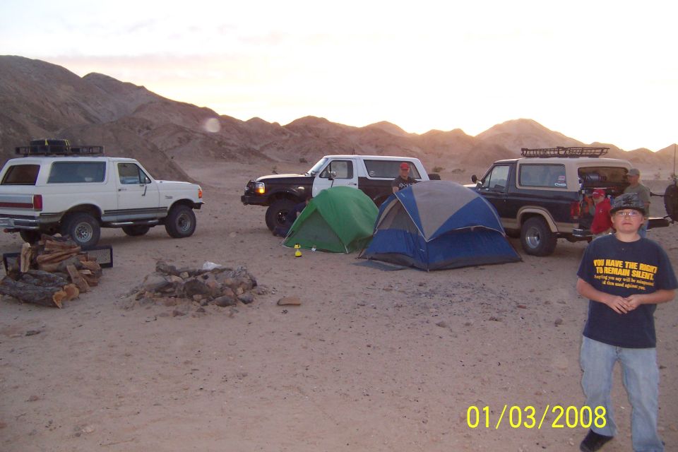 Three Ford Broncos parked in desert with tents and a campfire. A boy stands in the foreground