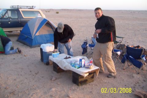 1990 Ford Bronco - A group is gathered around a table with food and coolers at a desert campsite, with a Ford Bronco parked nearby.