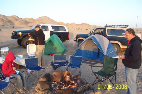 1990 Ford Bronco - Two Ford Broncos are parked at a desert campsite, with tents and a campfire set up.