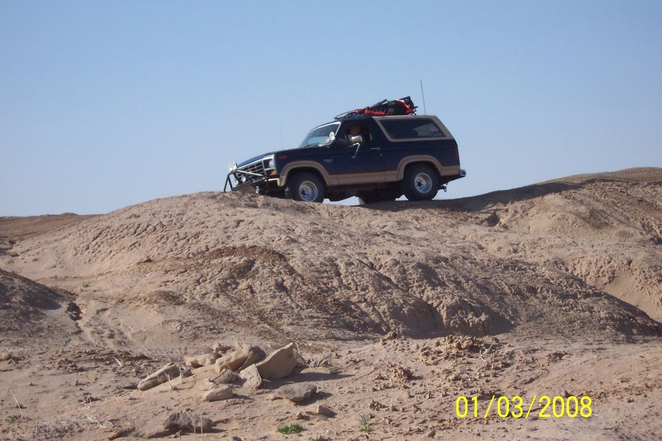 Dark blue and tan Ford Bronco with roof rack and brush guard on sandy hill