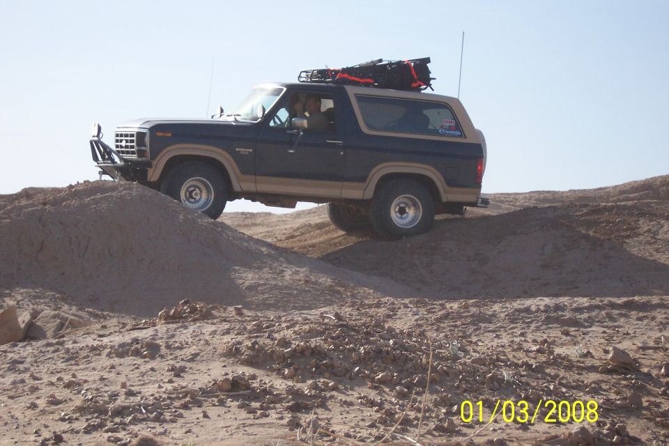 Dark blue and tan Ford Bronco climbing a dirt hill, roof rack loaded with gear