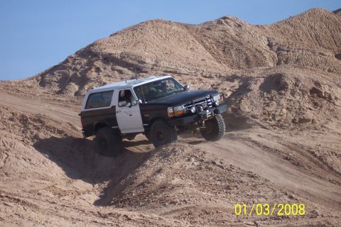 1990 Ford Bronco - A two-tone Ford Bronco navigates a sandy incline under a clear blue sky.