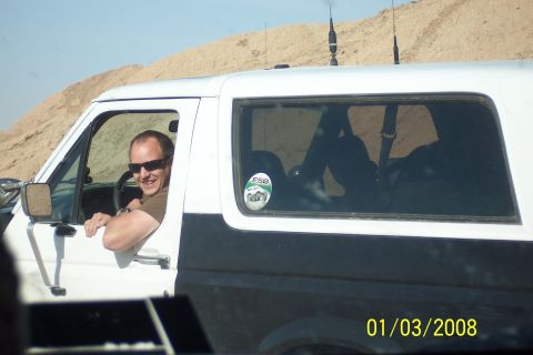 1990 Ford Bronco - A man smiles from the driver's seat of a two-tone Ford Bronco, with a sandy hill in the background.