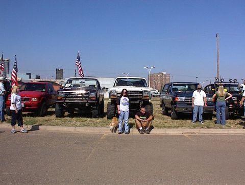 1981 Ford Bronco - A group of people gather around several trucks and SUVs parked outdoors under a clear blue sky.