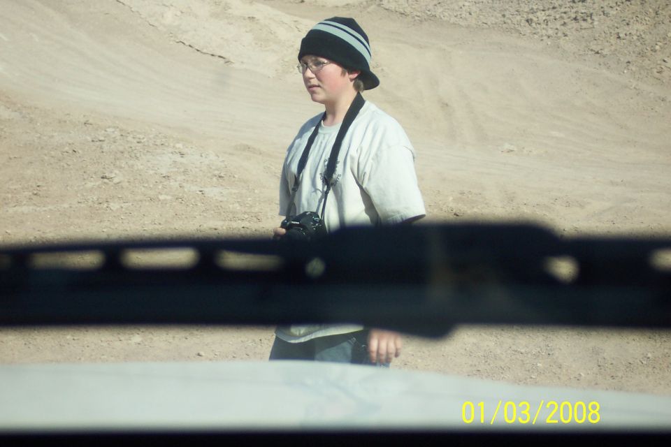 Person wearing a beanie and glasses, holding a camera, stands in a desert landscape
