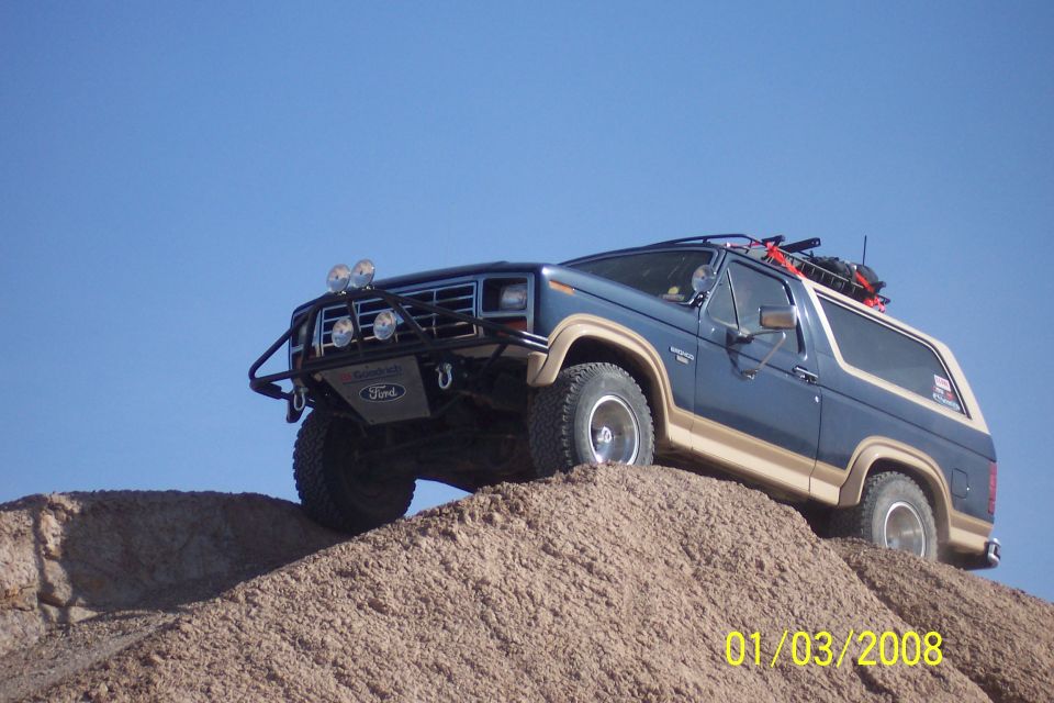 Blue and tan Ford Bronco with off-road lights and roof rack climbing a dirt mound against a clear blue sky