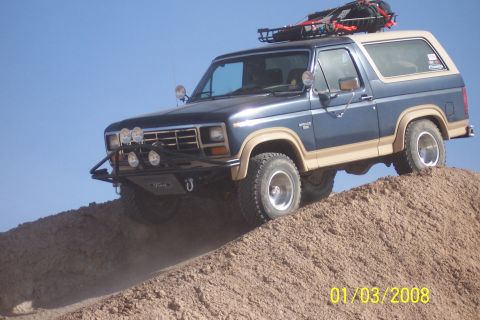 1990 Ford Bronco - A modified Ford Bronco navigates an off-road incline under a clear blue sky.