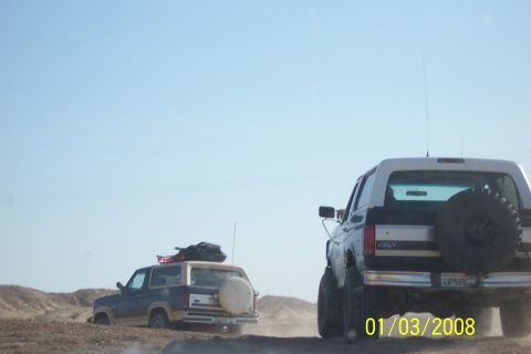 1990 Ford Bronco - Two Ford Broncos traverse a dusty, off-road path under a clear blue sky.