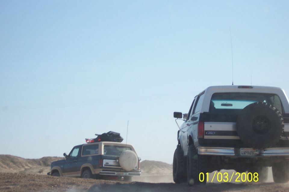 Rear view of a 1990 Ford Bronco with a spare tire on the back, driving on a dirt road