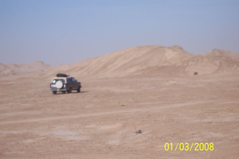 SUV with spare tire on back driving in a sandy desert with hills in background