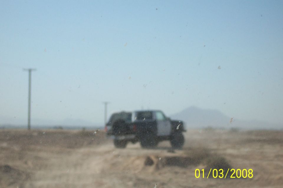 Dark SUV with spare tire on rear, driving on a dirt road under a clear sky