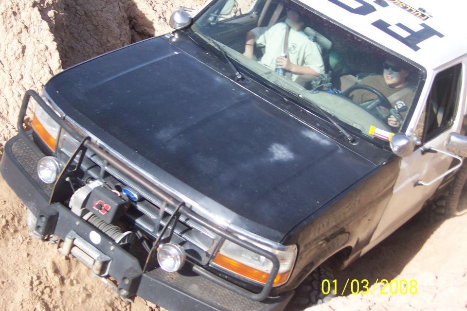 1990 Ford Bronco with winch and brush guard driving off-road, two people inside