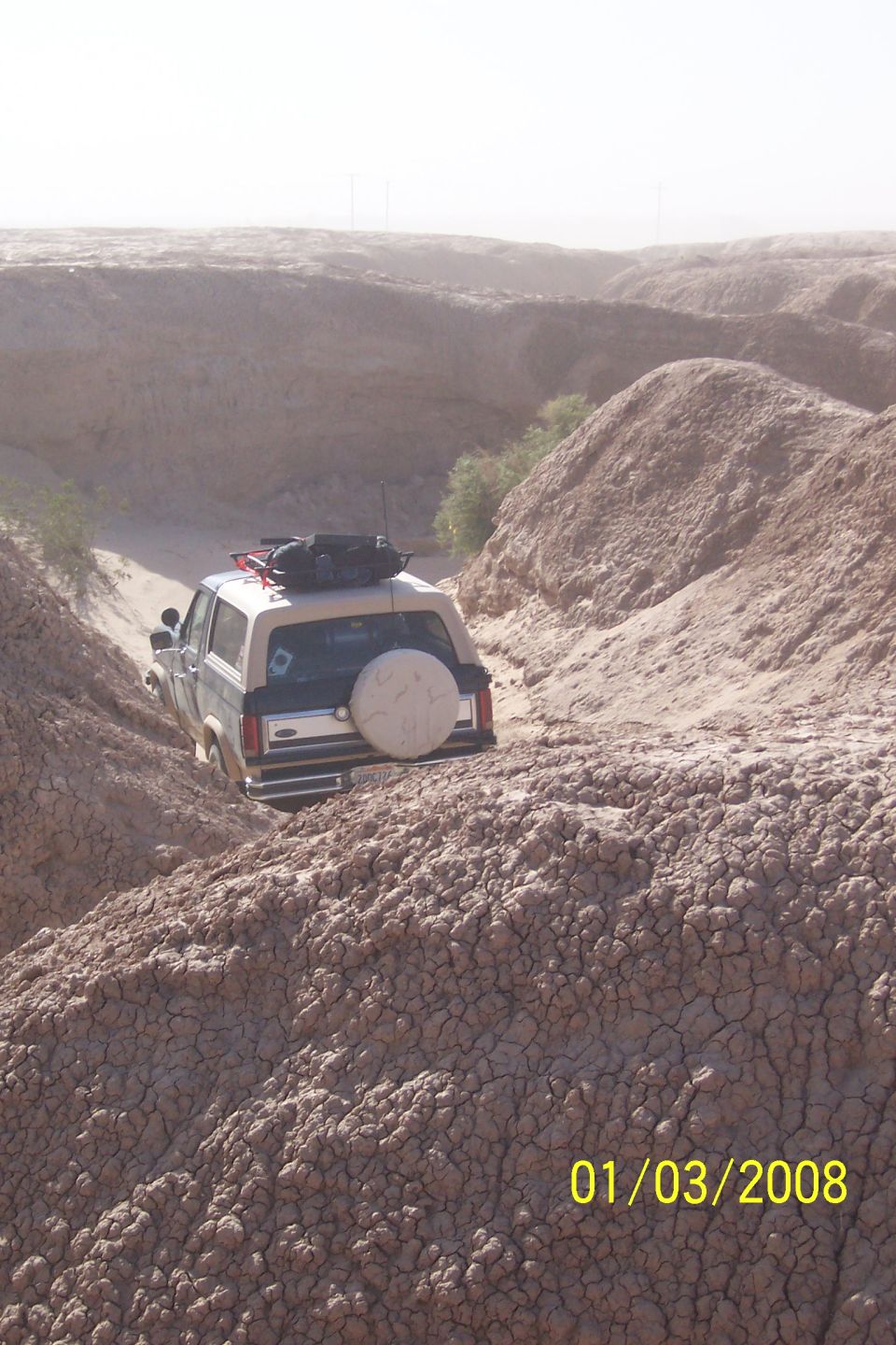 Ford Bronco driving down a dirt path in a dry, eroded canyon. It has a spare tire on the back and gear on the roof