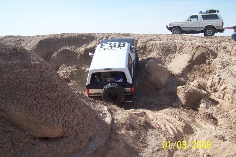 1990 Ford Bronco - Two Ford Broncos traverse a challenging sandy terrain under a clear sky.