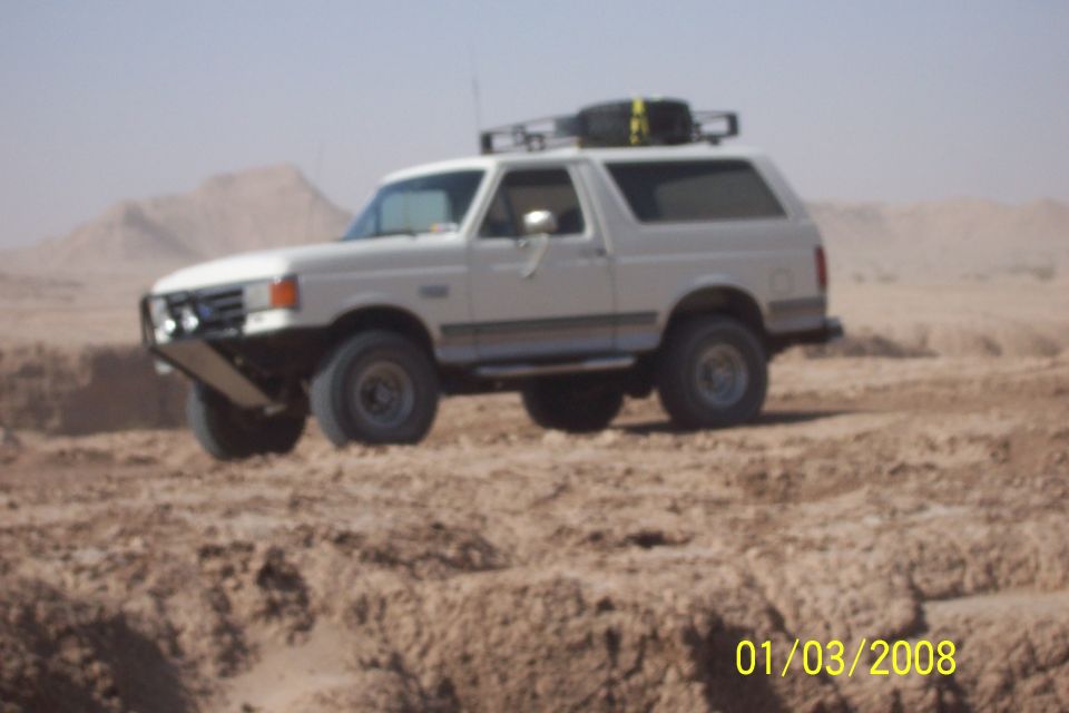 White 1990 Ford Bronco with aftermarket bumper and roof rack driving on a dirt mound