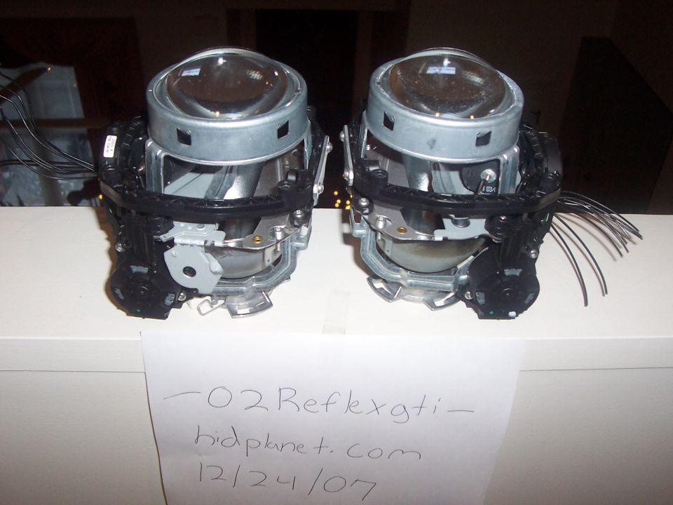 Two automotive projector headlights on a white surface with handwritten text below.