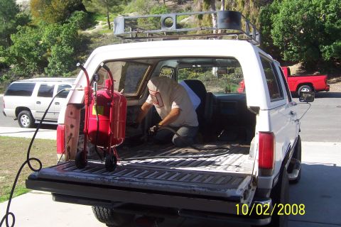 1990 Ford Bronco - A person in a protective suit is sandblasting the interior of a white Ford Bronco with the tailgate open. A red sandblaster tank is positio…