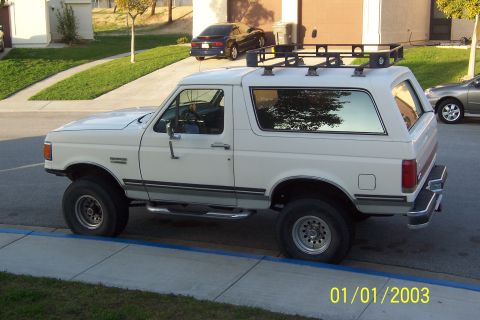 1990 Ford Bronco - The side view of a white 1990 Ford Bronco is parked on a suburban street next to a sidewalk. It has a roof rack and aftermarket wheels.