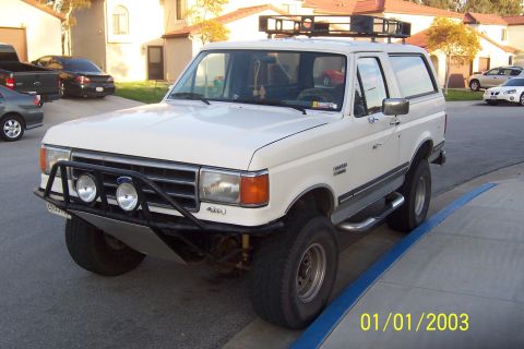 1990 Ford Bronco - A white 1990 Ford Bronco with a custom front bumper and roof rack is parked on a residential street next to a blue curb. The vehicle has a…