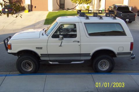 1990 Ford Bronco - A white 1990 Ford Bronco is parked on the side of a residential street. The SUV has a roof rack and a brush guard on the front.
