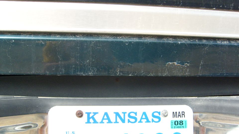 Scratched teal truck tailgate above chrome bumper with Kansas plate, MAR 08 tag