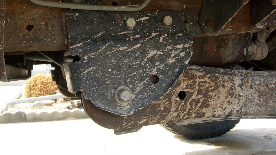 Underbody shot of a black skid plate bolted to a rusty truck frame rail with paint spatter