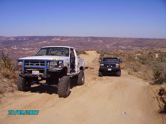 Lifted white Ford pickup truck with blue brush guard and black Ford pickup truck on dirt road.