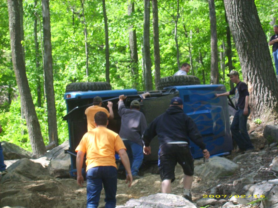 People trying to flip over a blue off-road vehicle in a forest.