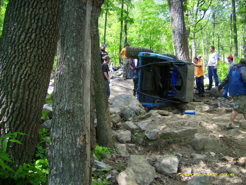 Overturned blue off-road vehicle on a rocky trail with people around.