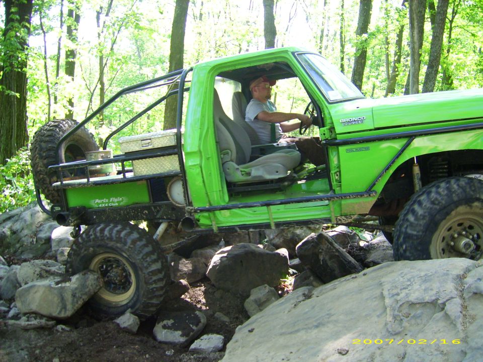 Green custom Ford Bronco off-roading over rocks with a roll cage and large tires.