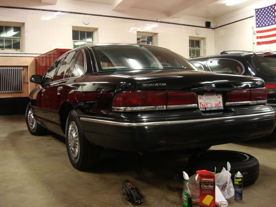 Black Ford Crown Victoria parked indoors, rear passenger side view, with cleaning supplies and tire on floor.