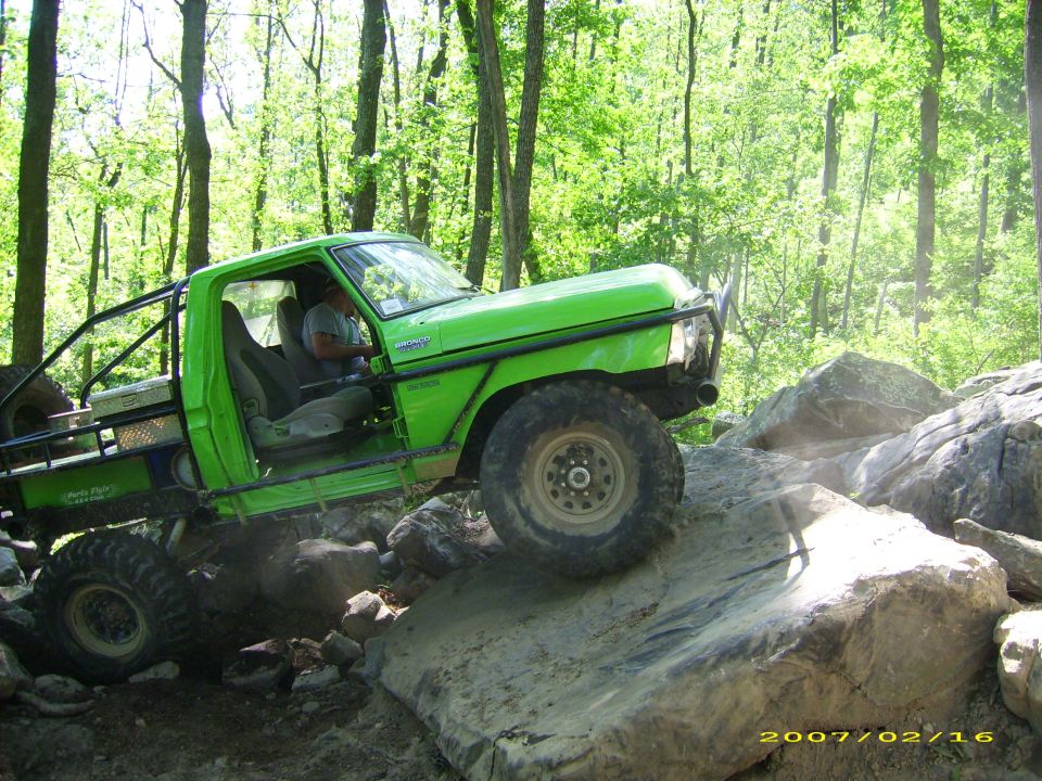 Green custom off-road Bronco climbing a large rock on a wooded trail.