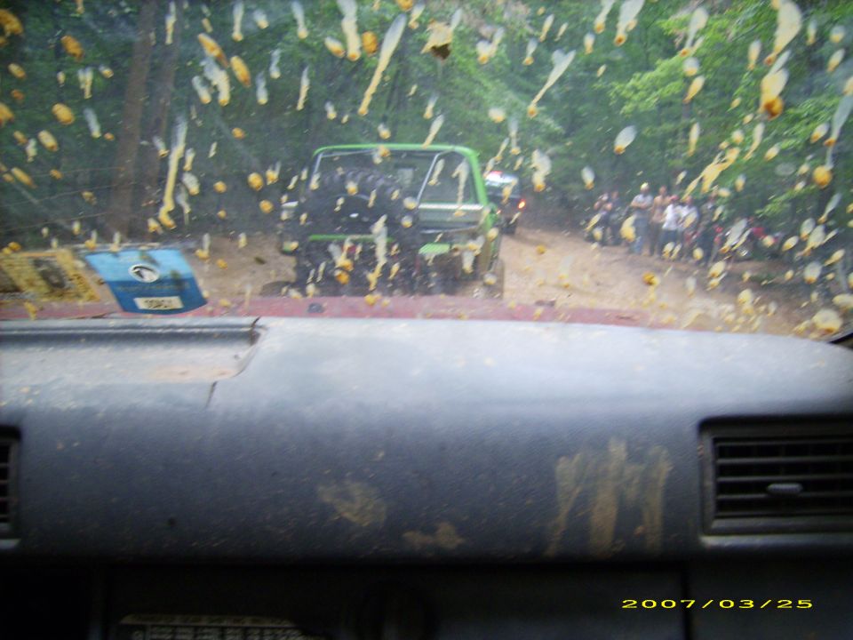 View through a dirty windshield of a green SUV on a dirt road with people. Dashboard in foreground.