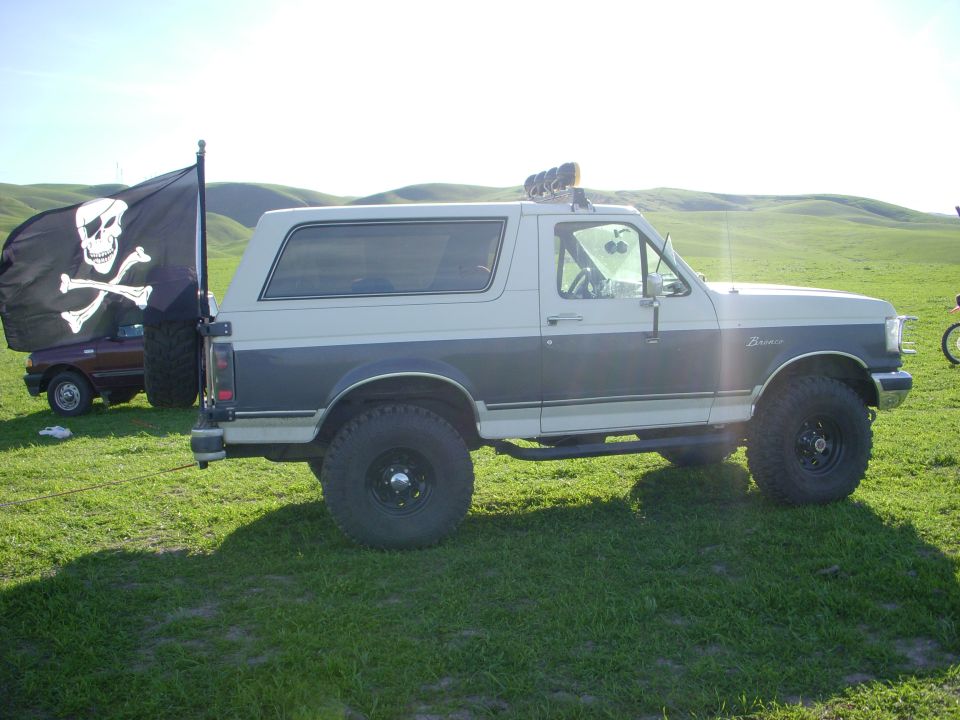 Lifted white and gray Ford Bronco parked on green hills beside a Jolly Roger flag