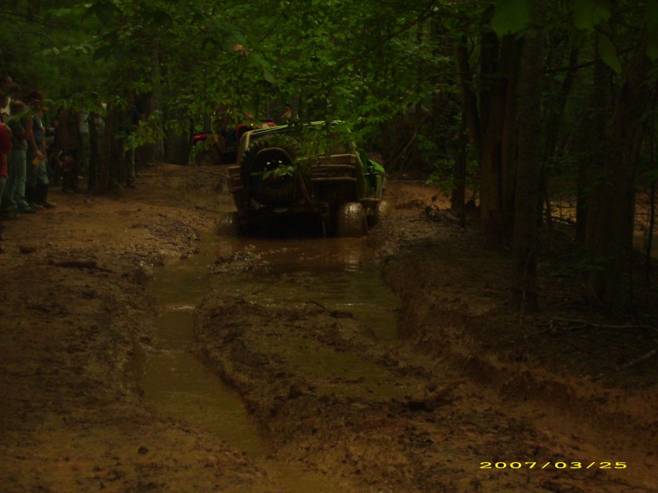 Green off-road vehicle driving through a muddy rut in a forest.