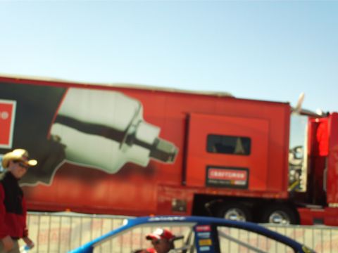 1978 Ford Bronco - A large red trailer with a graphic of a tool is parked under a clear blue sky. A blue race car is partially visible in the foreground.