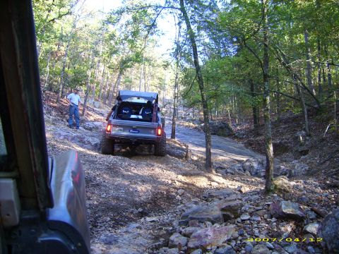 1978 Ford Bronco - A 1978 Ford Bronco navigates a challenging off-road path through a forest, with its cargo area open.
