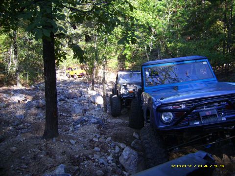 1978 Ford Bronco - Multiple off-road vehicles, including a 1978 Ford Bronco, traverse a challenging rocky path through a wooded area.
