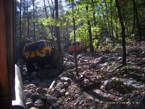 1978 Ford Bronco - Two off-road vehicles navigate a challenging rocky path through a dense forest.