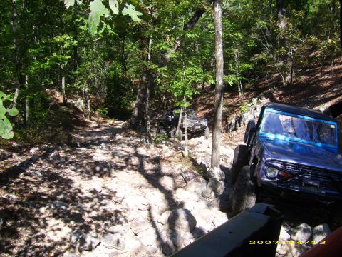 1978 Ford Bronco - Two off-road vehicles, including a 1978 Ford Bronco, tackle a challenging rocky trail through a wooded area.