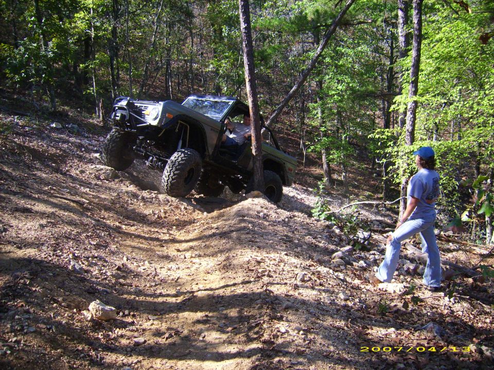 Off-road vehicle climbing a steep, wooded trail. Person watches from the side
