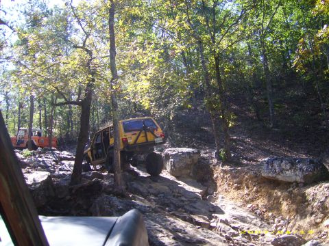 1978 Ford Bronco - Two off-road vehicles tackle a challenging rocky path through a wooded area.